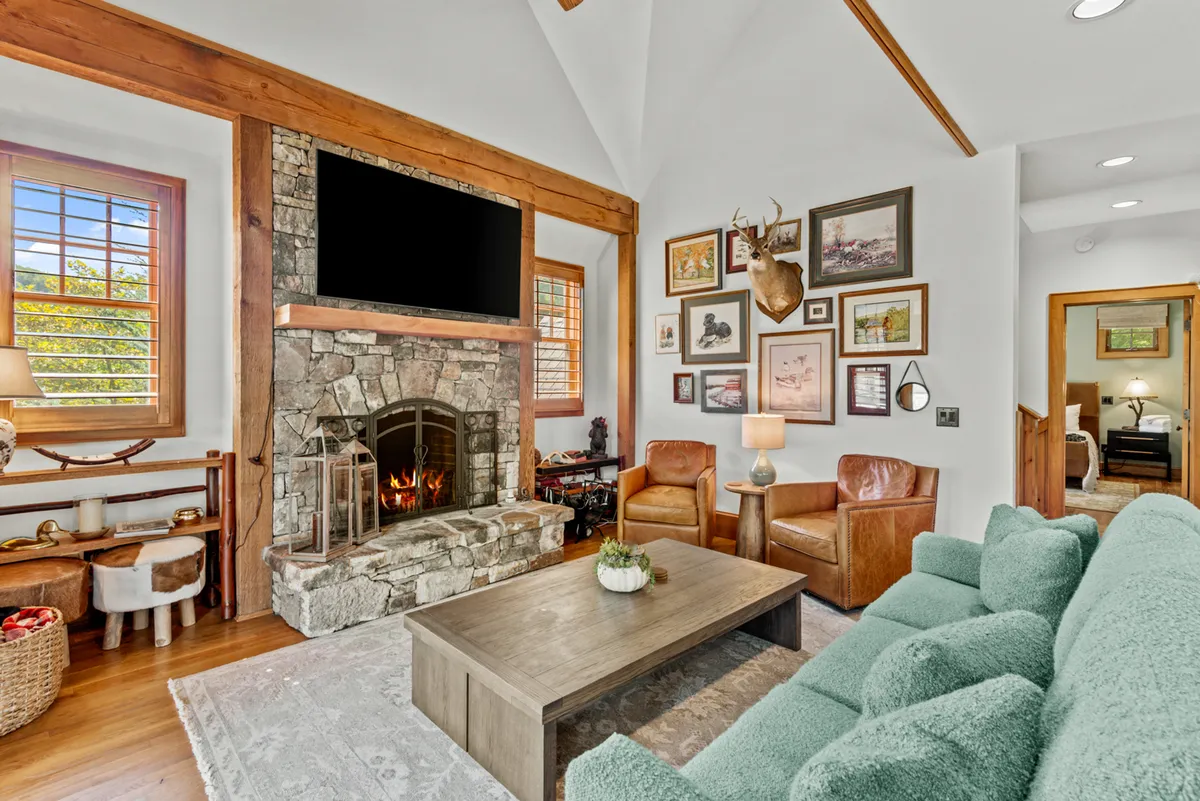 A spacious living room filled with warm, natural light, featuring a stone fireplace as its centerpiece. The room is furnished with a large green couch, comfortable armchairs, and accented by wooden beams and artwork on the walls. The arrangement is designed for relaxation and group gatherings, with an intimate, inviting atmosphere.