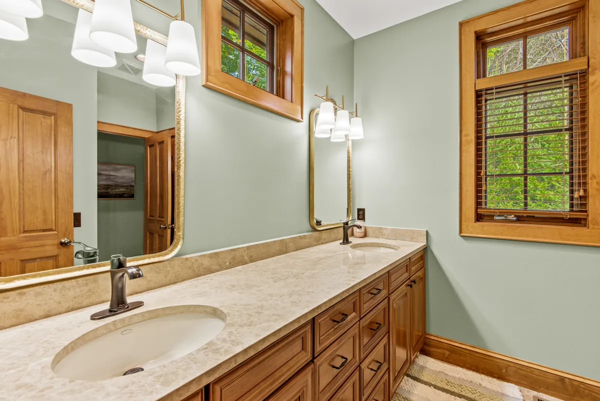 A spacious, light-filled bathroom featuring a double sink vanity, a large mirror, and windows allowing natural light to brighten the space. The clean, modern fixtures highlight both function and relaxation.