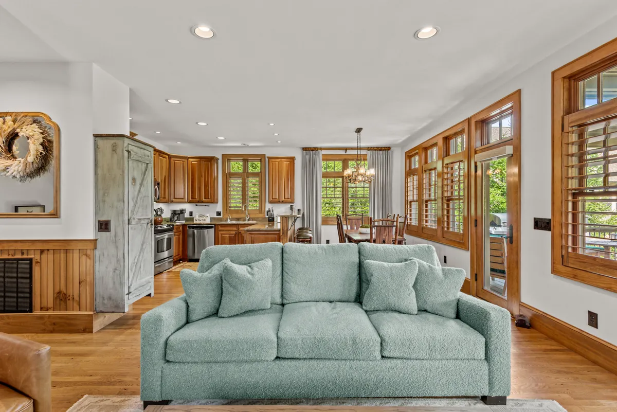 A bright and inviting open-concept space featuring a soft turquoise couch, wooden kitchen cabinets, and a dining area with wooden chairs. Large windows flood the room with natural light, while the kitchen and dining areas are seamlessly connected, ideal for both cooking and gathering. The scene suggests an environment where family and friends can easily flow from preparing meals to sharing them together.