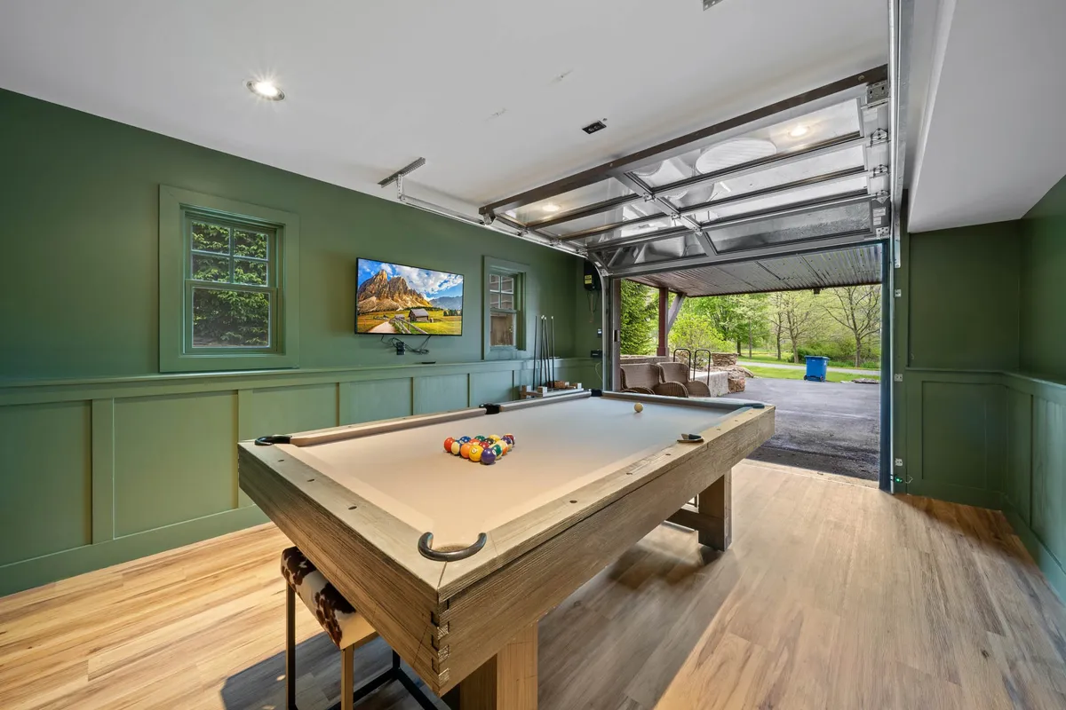 A lively interior space featuring a pool table set up for play, a television mounted on the wall, and a large garage door that opens to the outdoors. The room has ample space for games, watching TV, and socializing, with plenty of natural light streaming in from the open door.