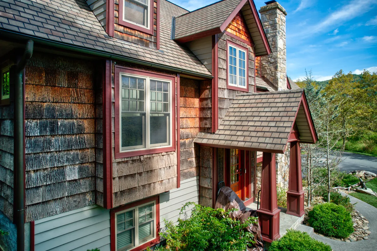 A cozy and inviting exterior view of a home with a wooden shingle facade, large windows, a stone chimney, and lush greenery. The porch area suggests relaxation and the opportunity for group gatherings, surrounded by nature.