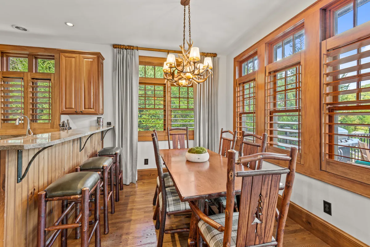 A warm and inviting dining room scene featuring a central dining table under a stylish chandelier, surrounded by windows that let in natural light. A bar counter is visible, offering flexibility for casual meals or festive gatherings. The overall ambiance is ideal for group dining and celebratory meals.