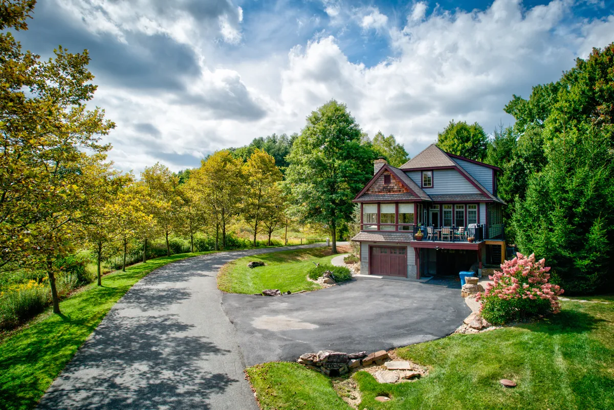 The home’s exterior is showcased with a well-kept garden, a long driveway, and mature trees providing privacy. The porch area hints at possibilities for relaxing with coffee or watching the sunset, while the overall setting conveys a sense of safety and tranquility within the gated community.