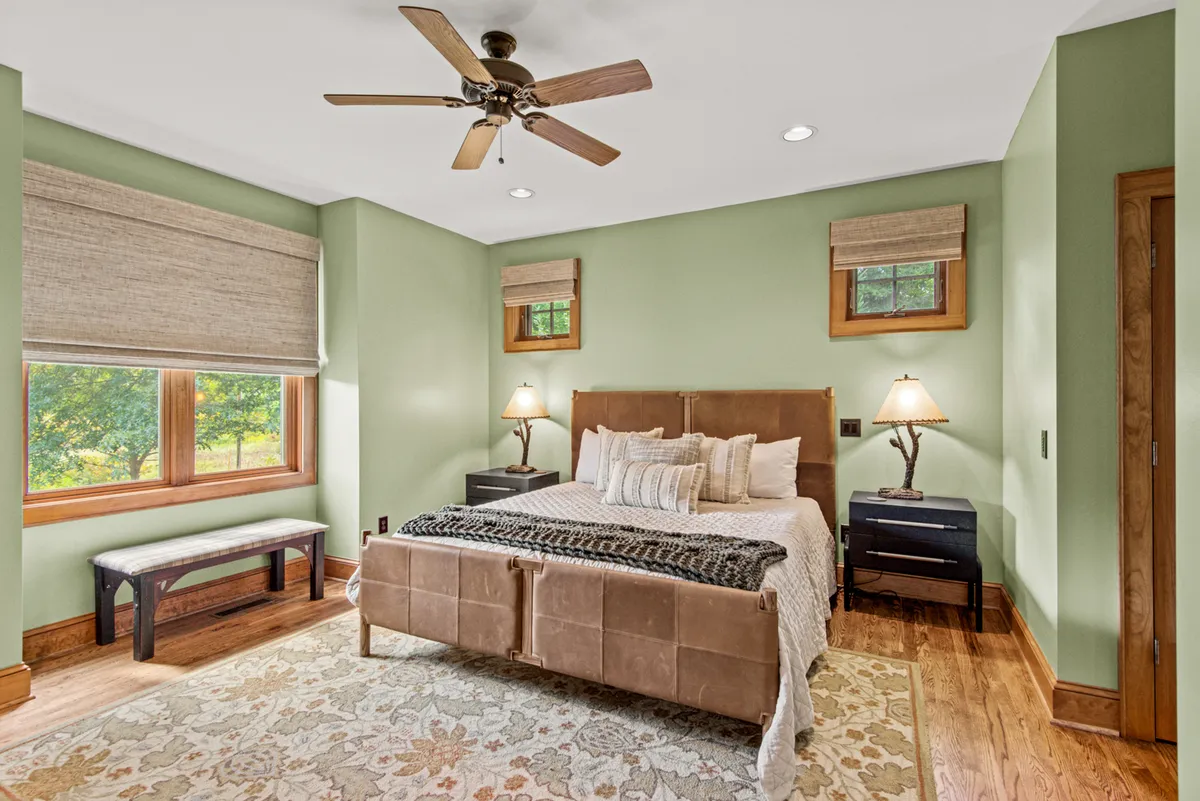 A serene bedroom featuring a large bed with decorative pillows, wooden nightstands, and expansive windows letting in soft natural light. The ceiling fan overhead and calming decor create an inviting, restful atmosphere perfect for winding down.