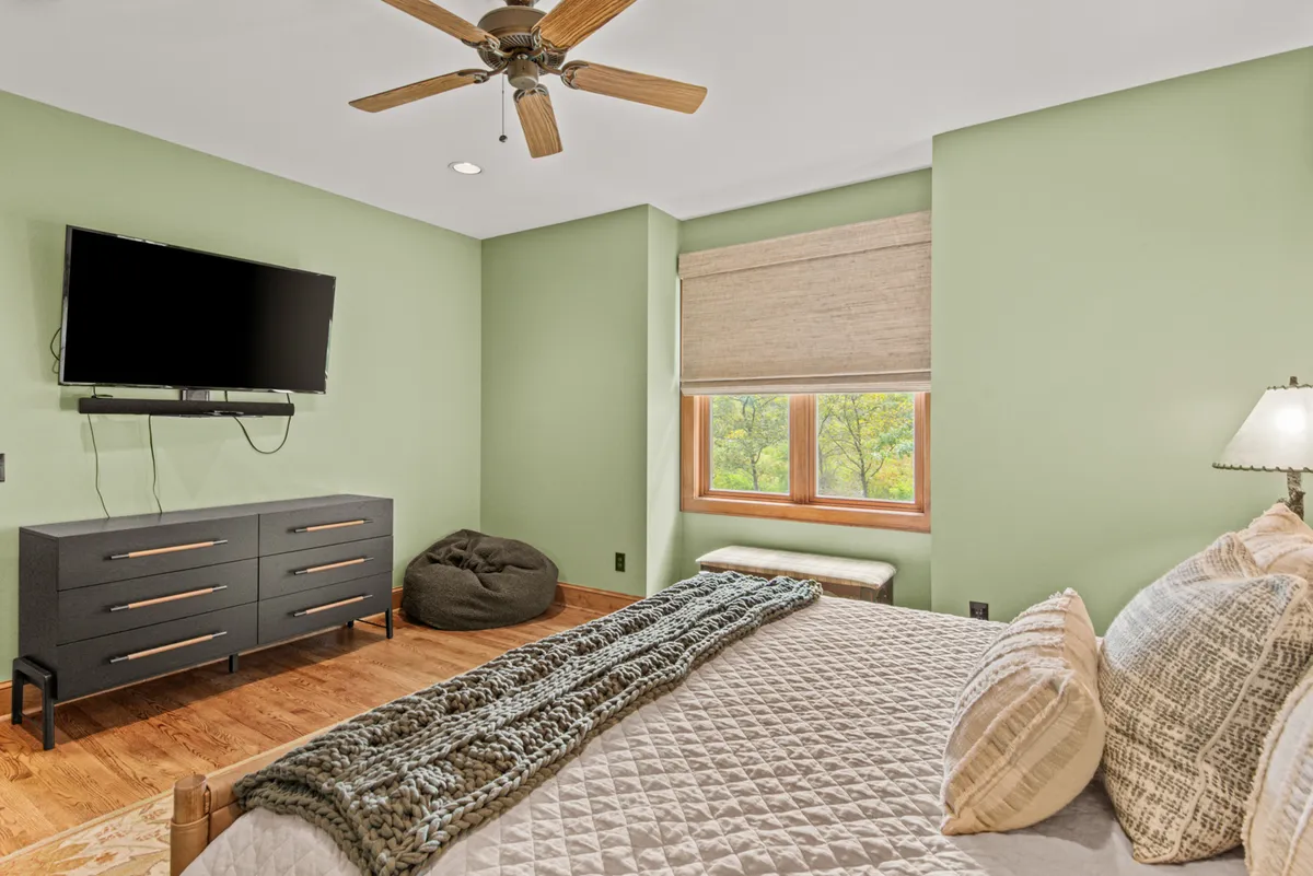 A cozy bedroom with a plush bed dressed in textured blankets and pillows, a wall-mounted television, and a window flooding the space with natural light. A bean bag chair adds a casual, comfortable touch ideal for both adults and kids.