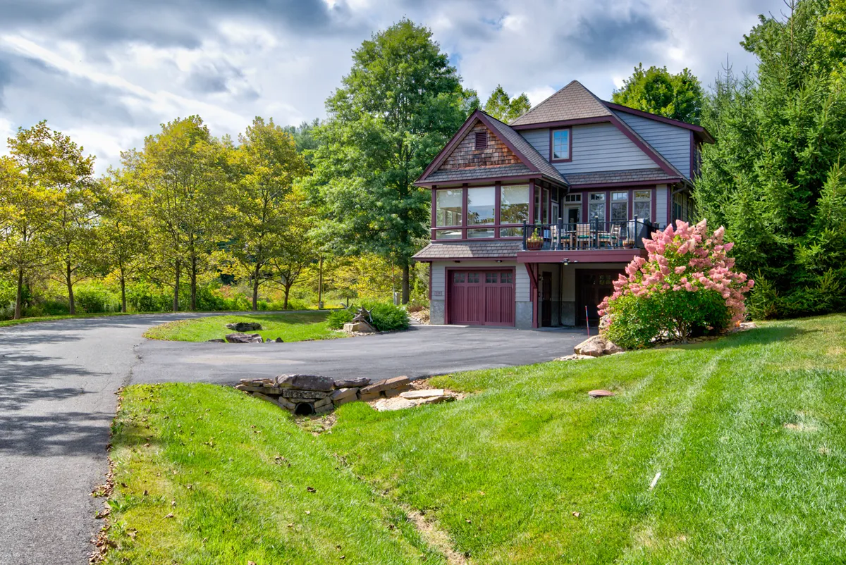 The image showcases the house surrounded by lush greenery and a flowering bush, conveying a sense of serenity and natural beauty. The porch is visible, suggesting a welcoming outdoor space ideal for relaxing or watching sunsets. The expansive lawn in the foreground hints at ample room for pets and families to play or unwind.
